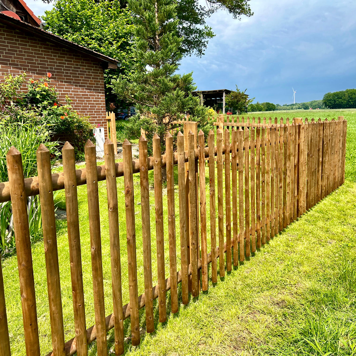 Langer Senkrechtzaun aus braunem Holz als Grundstücksgrenze an einer Wiese neben einem Haus